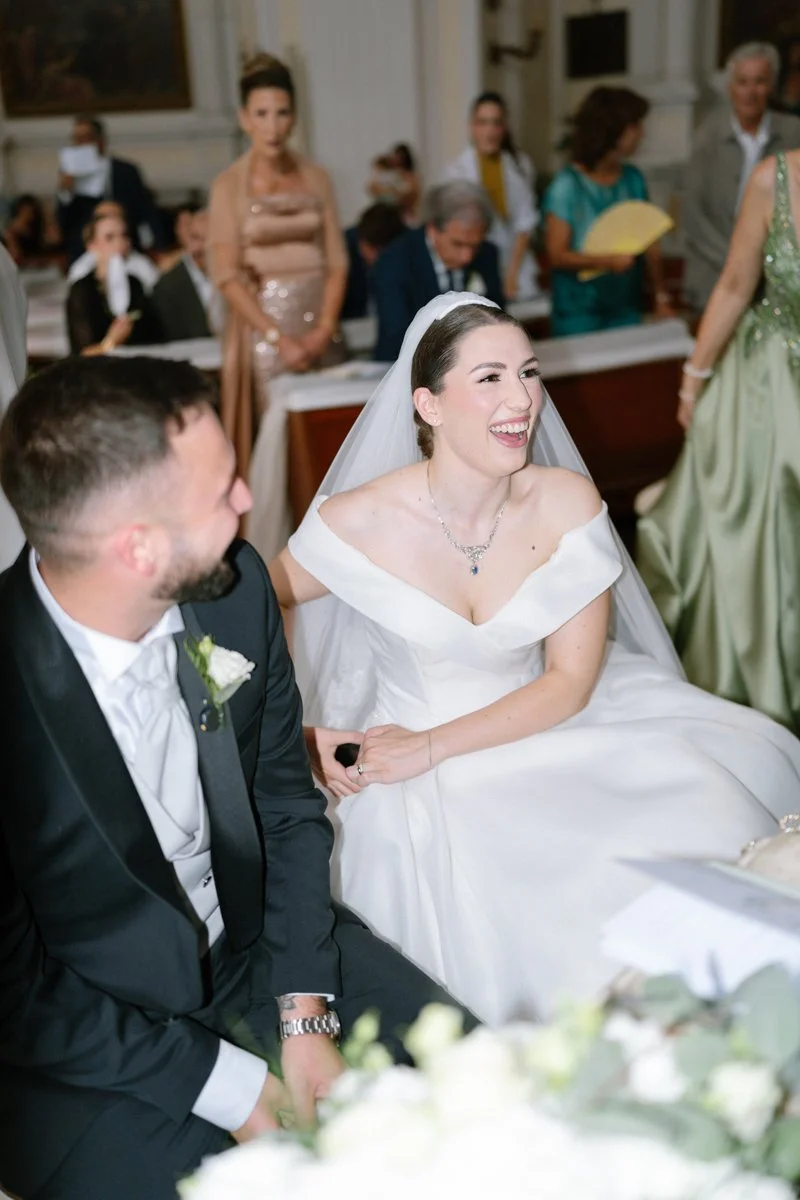 A bride and groom sitting together at their wedding reception, smiling and laughing. The bride is wearing a white wedding dress and veil, while the groom is in a dark suit. Guests are visible in the background, some standing and others seated, in a f