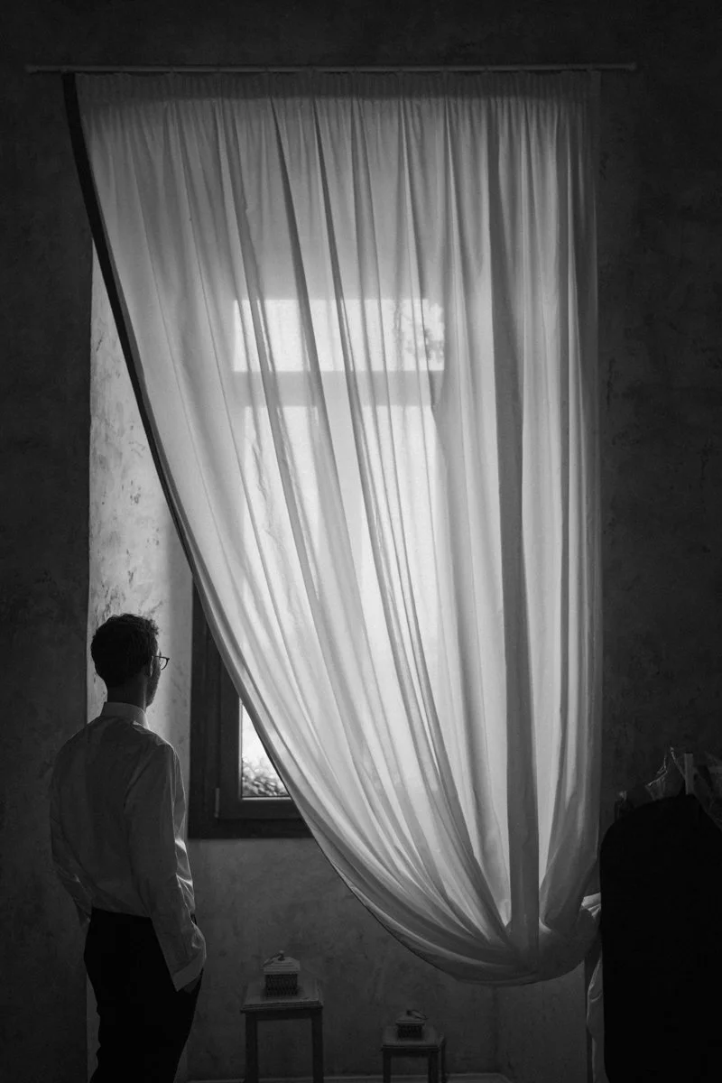 A man stands in a dimly lit room with a large window covered by flowing white curtains. Light filters through the curtains, illuminating the room with soft natural light.