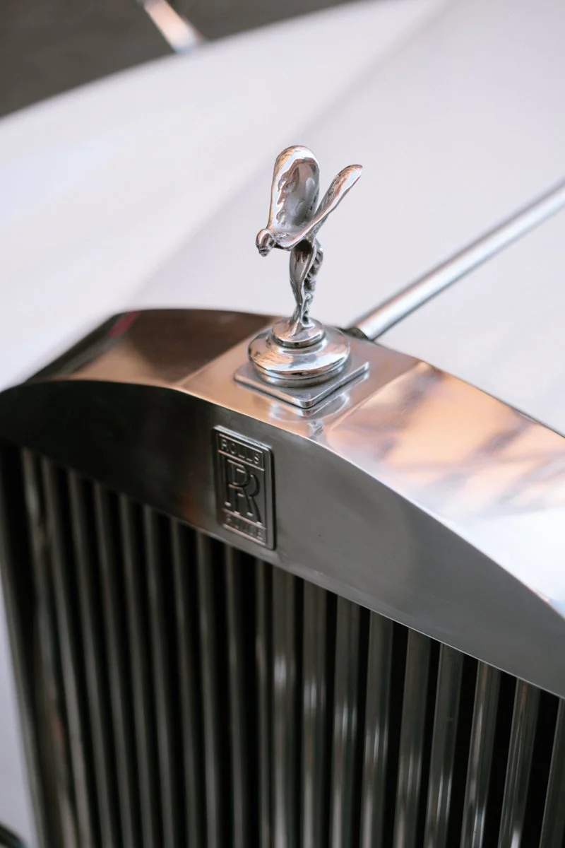 Close-up of a silver hood ornament shaped like a woman with wings, on a vintage car with a chrome grille and the Bentley logo.