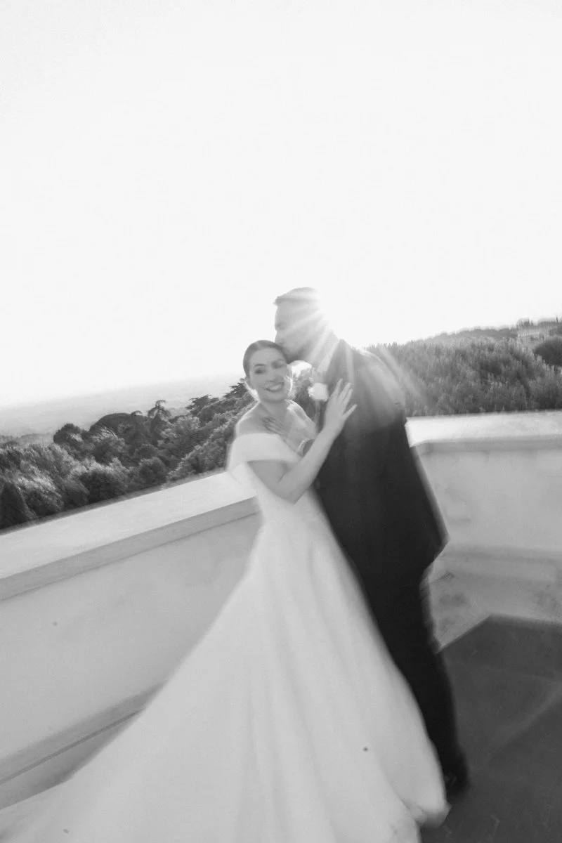 A bride and groom embrace on an outdoor balcony with a scenic landscape in the background, illuminated by bright sunlight.