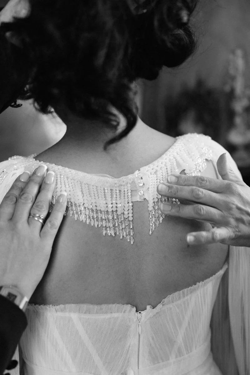 A woman with dark, curly hair is having a dress adjusted on her shoulders. The dress has an embellished beaded collar with dangling beads. The image shows her back and the hands of someone helping her, with a blurred background.