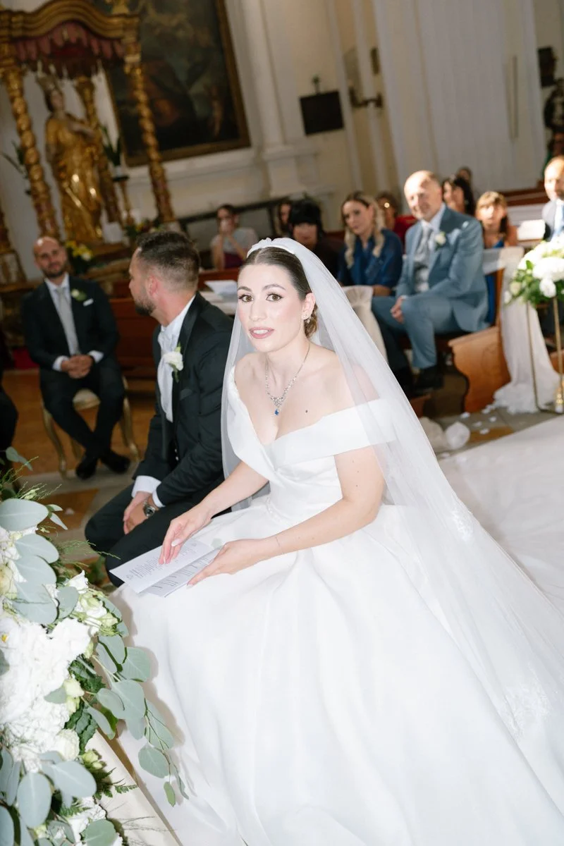 A bride in a white wedding gown and veil seated in a church, holding a paper, with guests seated behind her.
