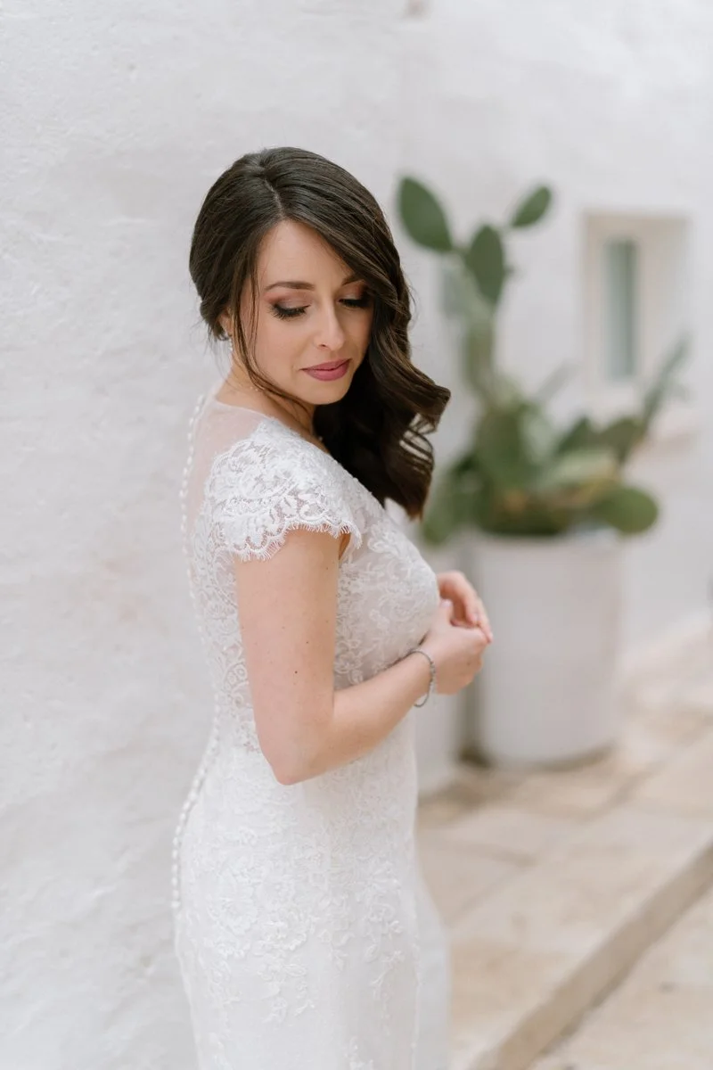 A woman wearing a white lace dress with dark, wavy hair standing against a white wall with a green potted plant in the background.