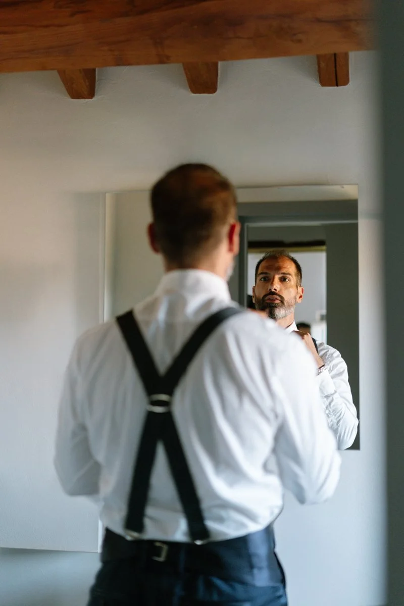 A man in a white shirt with black suspenders looking into a mirror and adjusting his collar.