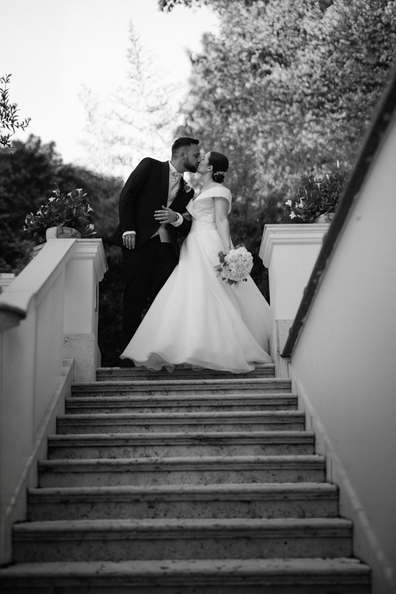A black and white photograph of a newlywed couple kissing on a staircase outdoors, with trees in the background. The bride wears a white wedding gown and holds a bouquet, while the groom is dressed in a dark suit.