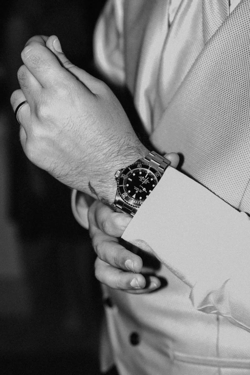 A close-up of a man's wrist showing a Rolex watch, with one hand clasped over the other, wearing a suit jacket and shirt.