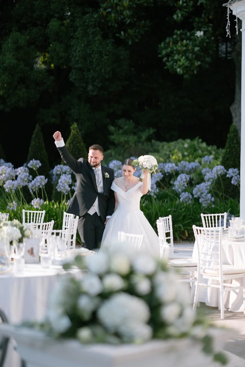 A bride and groom celebrating outdoors during their wedding reception, with the bride holding a bouquet and the groom raising his fist in celebration.