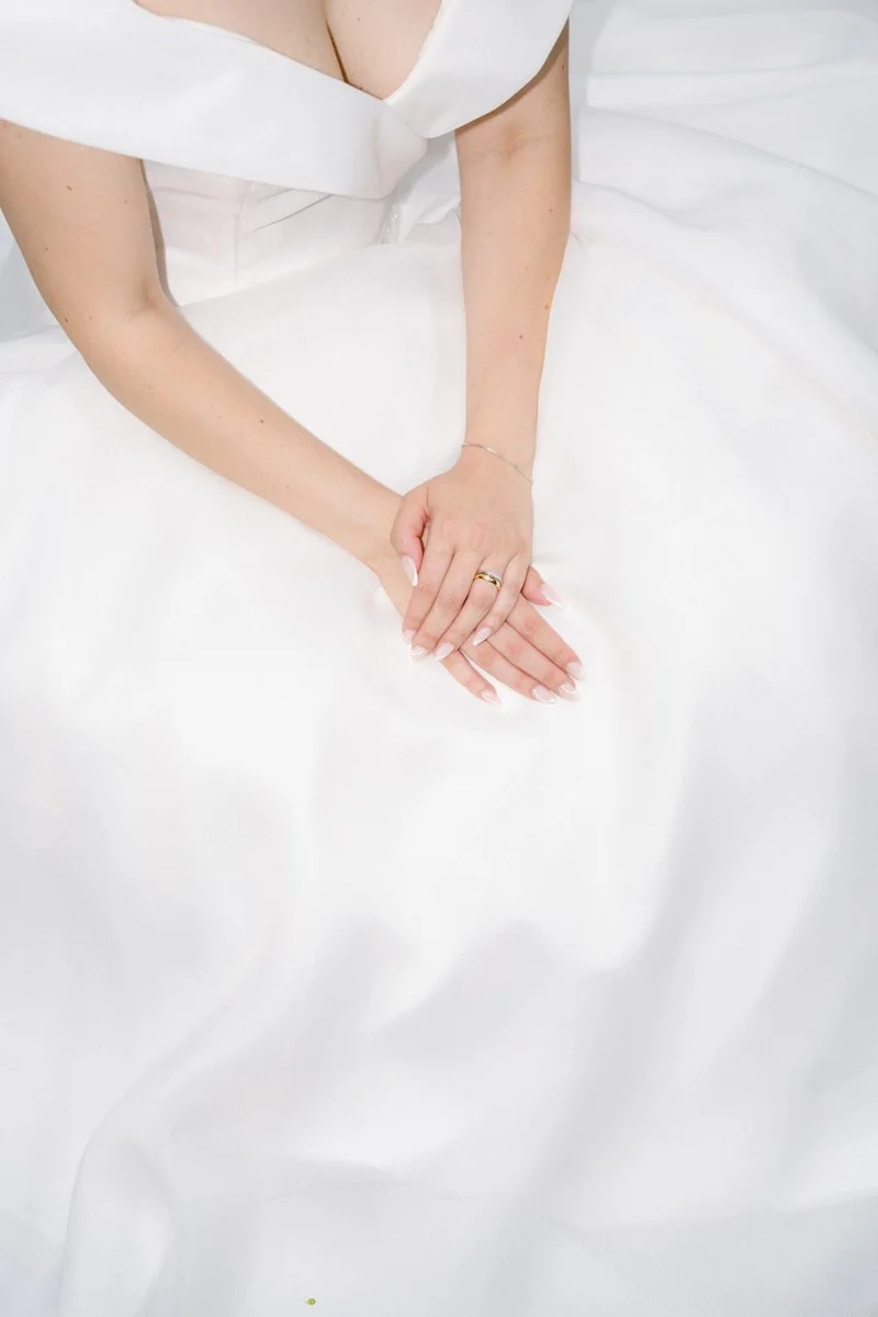 Close-up of a woman in a white wedding dress sitting with hands folded on her lap.