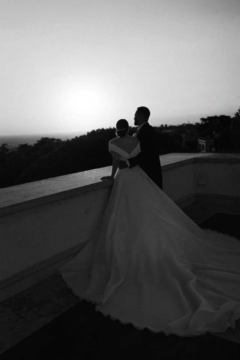 A couple dressed in wedding attire standing on a balcony, gazing at the sunset or sunrise over a landscape with trees.