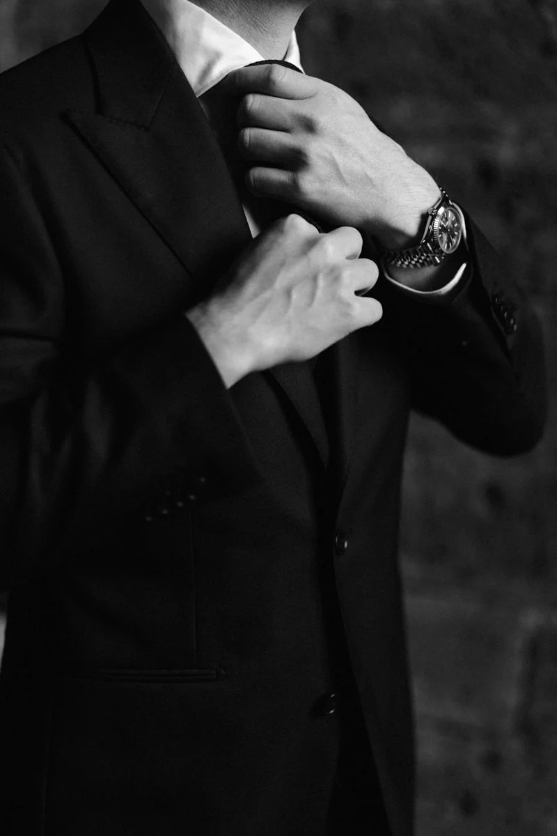 A man in a black suit adjusting his tie, wearing a wristwatch, in a black-and-white photograph.