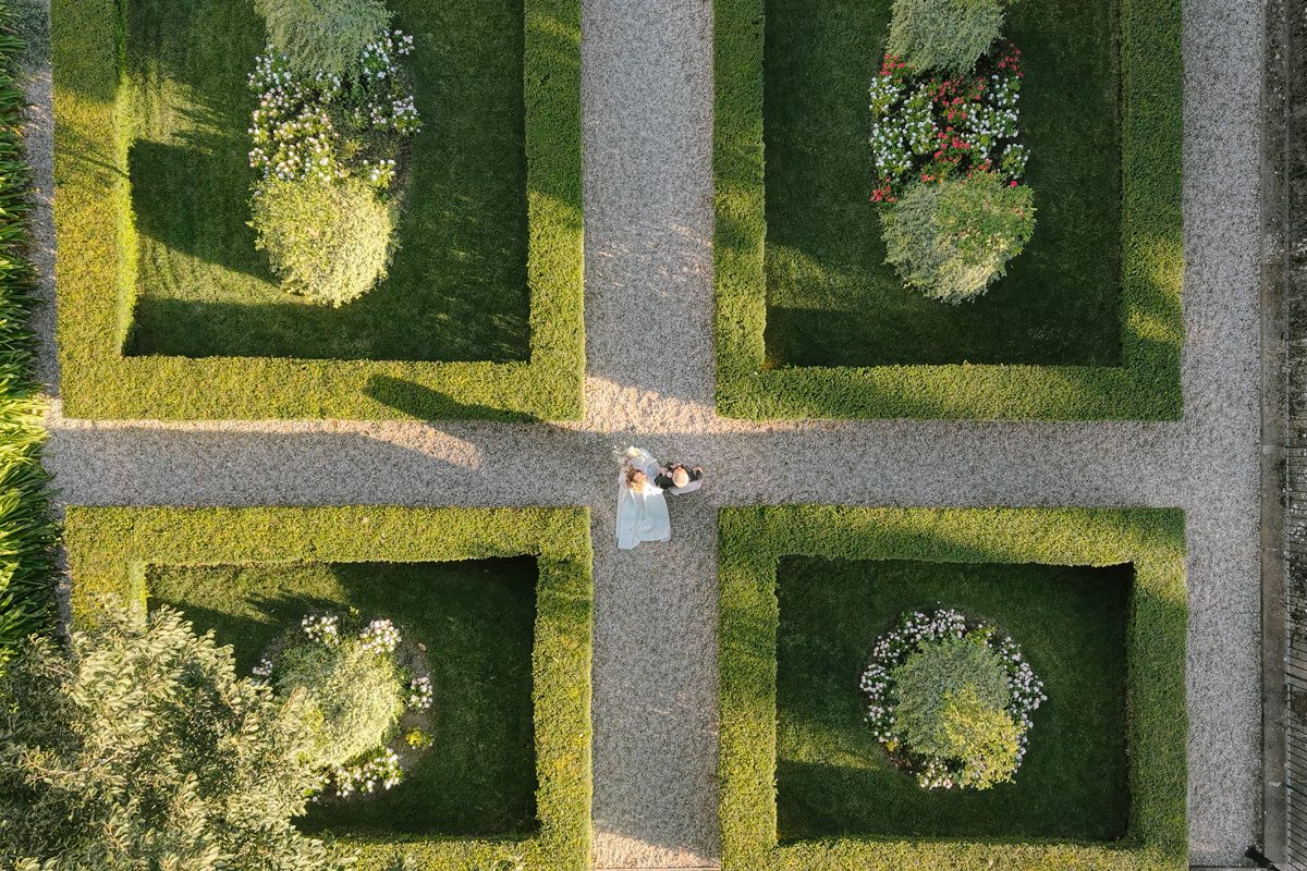 Top-down view of a bride and groom walking down a garden path surrounded by neatly trimmed hedges and flower beds.