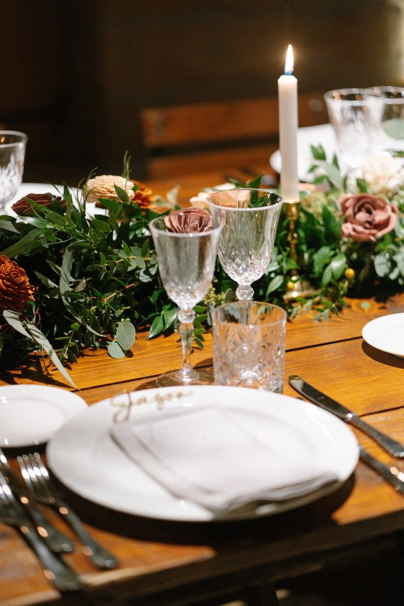 A dinner table set with white plates, silverware, crystal glasses, a lit white candle, and a floral centerpiece of greenery and pink, purple, and orange flowers.