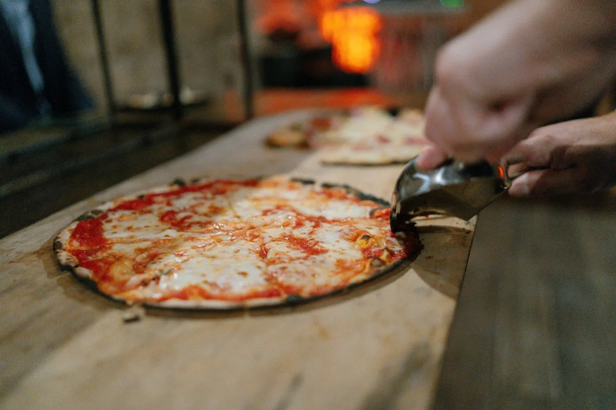 A person slicing a cheese pizza on a wooden surface inside a restaurant or pizzeria.