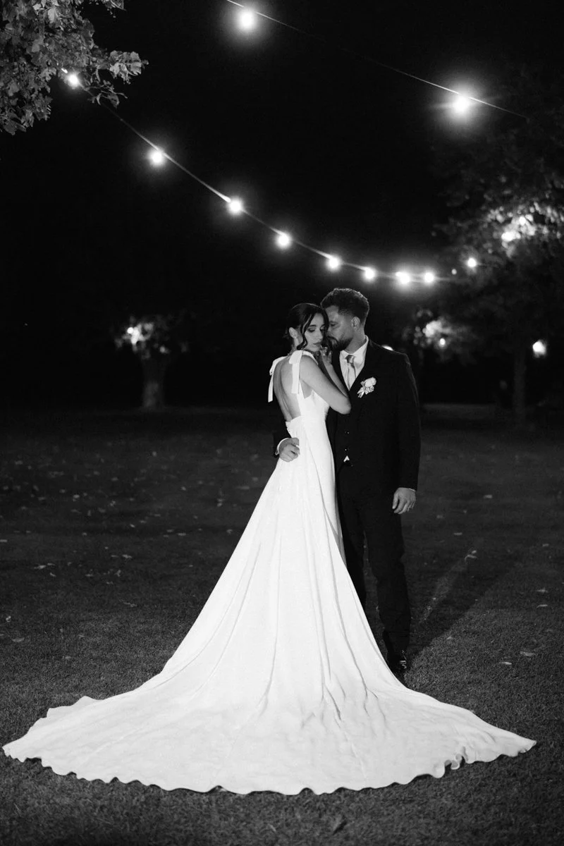 A bride and groom embrace under outdoor string lights at night, with trees in the background. The bride wears a long white gown, and the groom is in a dark suit.