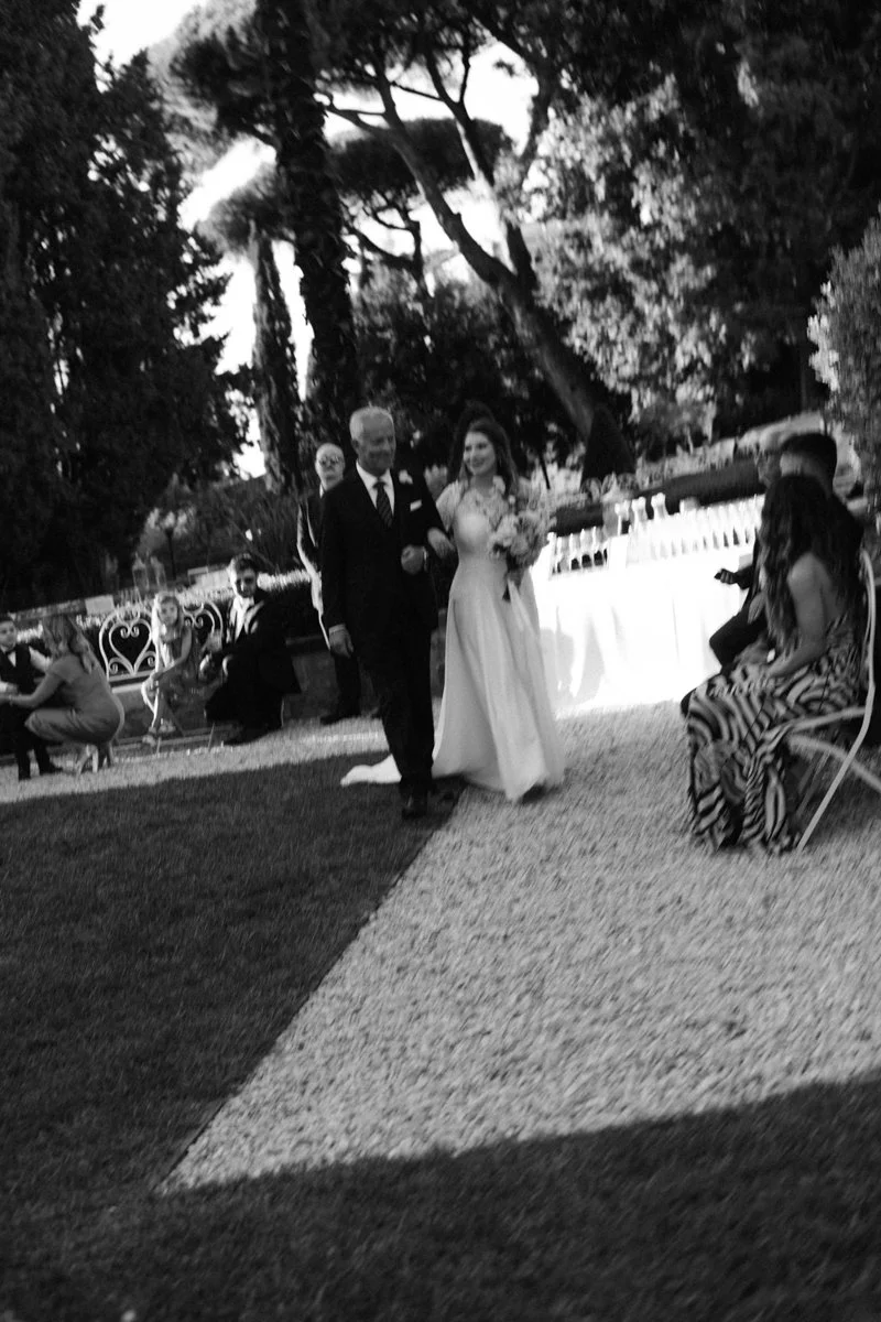 Black and white photo of a wedding ceremony outdoors, with a bride in a white gown walking arm-in-arm with a man in a suit, surrounded by seated guests and tall trees in the background.