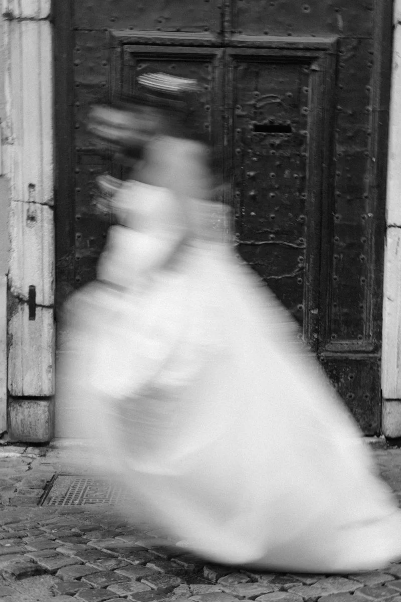 A blurry figure of a woman in a white dress walking past an old wooden door with stone walls and a cobblestone street.