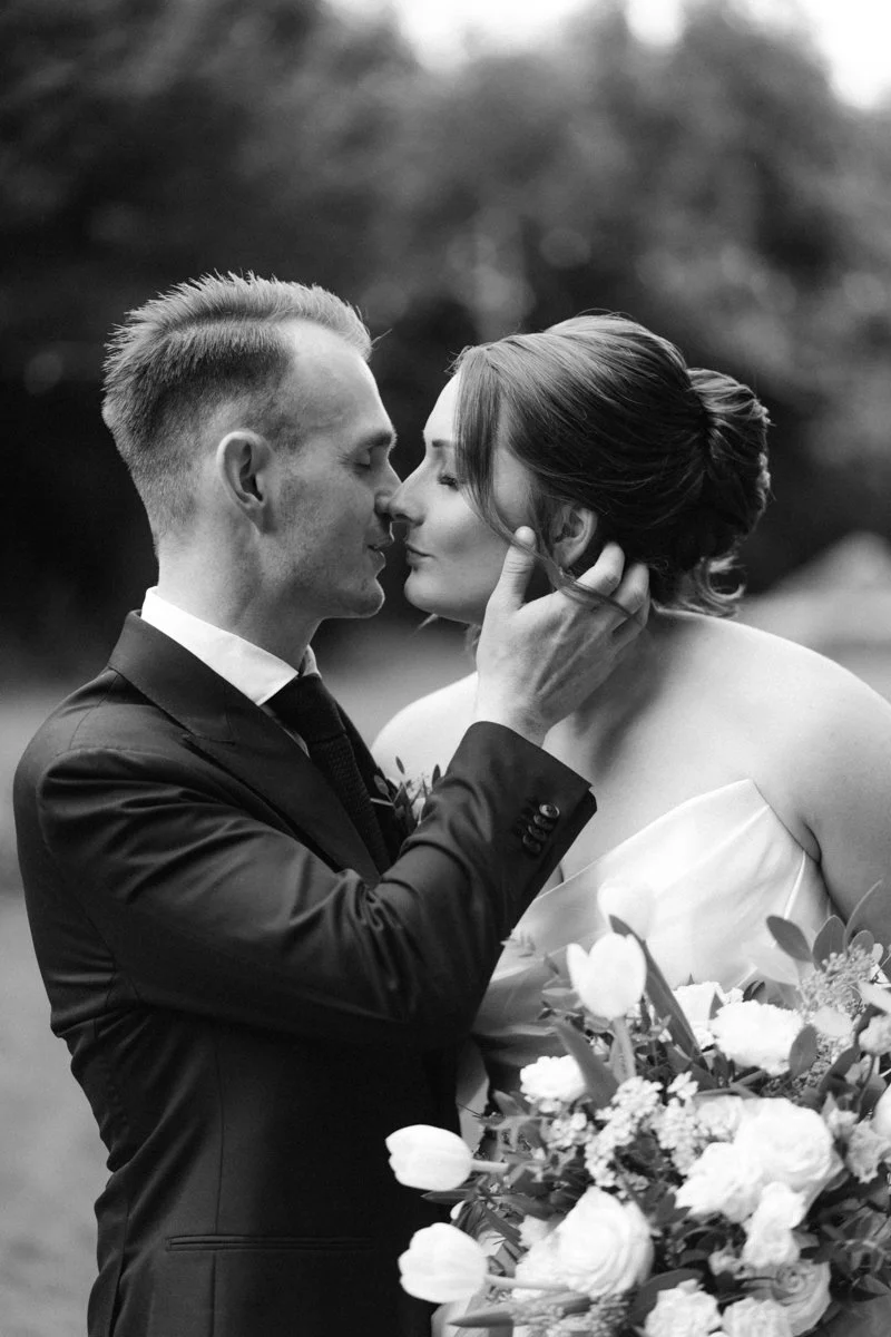 A black-and-white photo of a wedding couple, with the groom gently holding the bride's face as they about to kiss, with flowers in the bride's arms and a blurred outdoor background.