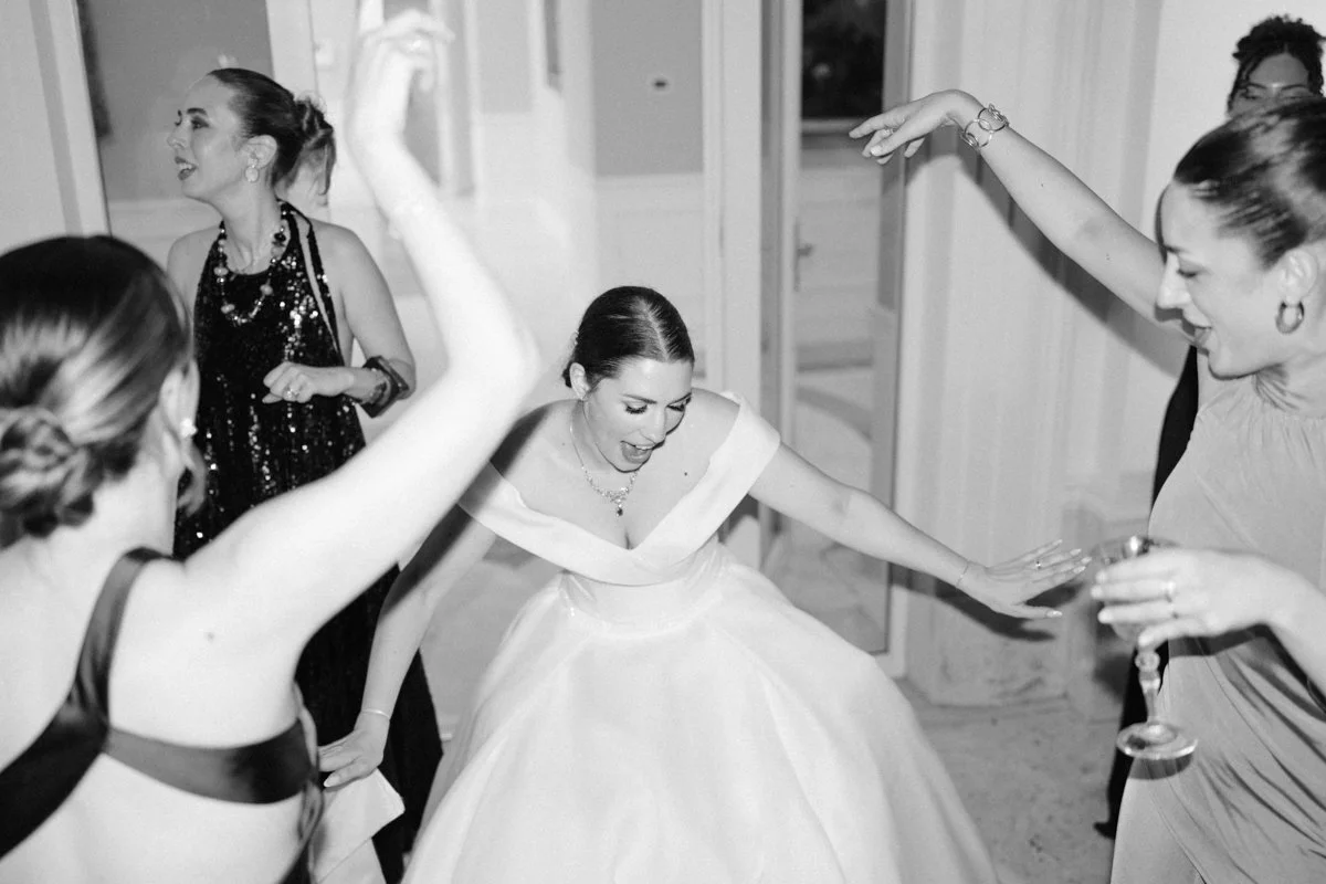 Women dancing and celebrating at a wedding reception, including a bride in a white gown, in a lively indoor setting.