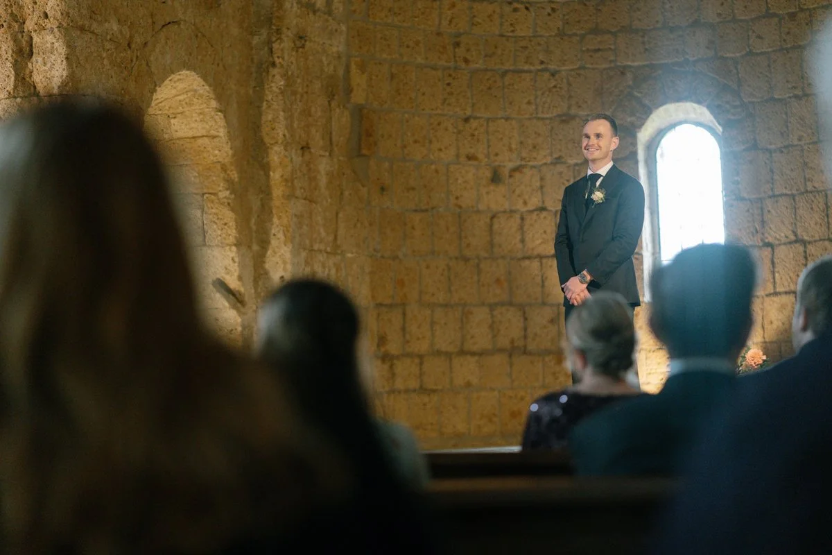 A groom in a black suit and tie stands smiling in a church on his wedding day.