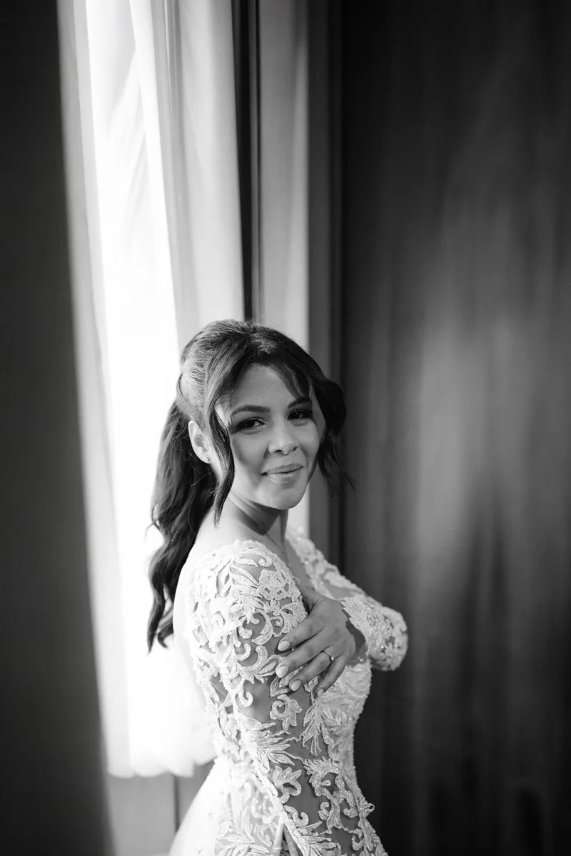 A woman in a wedding dress with lace detailing, standing near a window with curtains, smiling and looking at the camera.