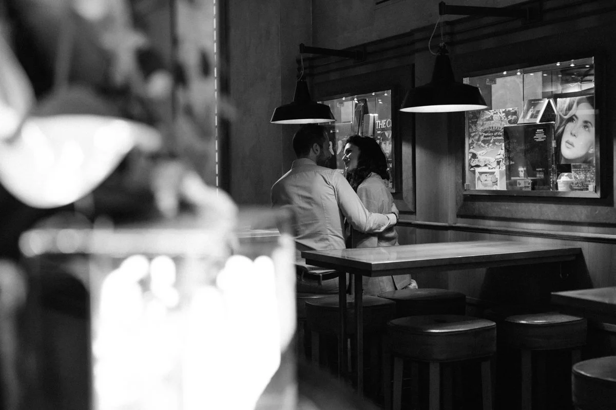 A black and white photo of a couple sitting close together at a restaurant table, smiling and looking at each other, with a window display of magazines and pictures behind them.