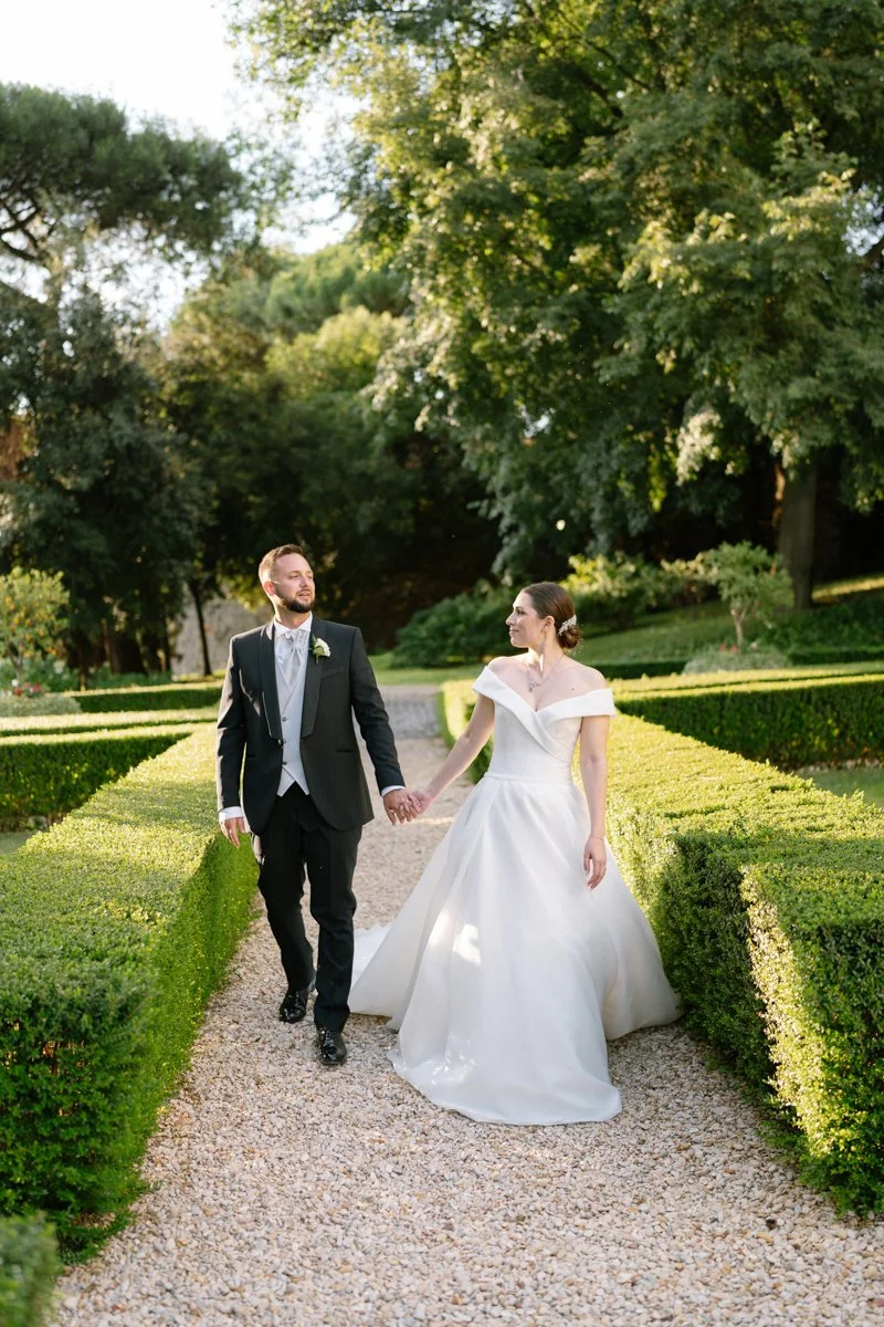 A bride and groom holding hands and walking in a garden with trimmed hedges and tall trees.