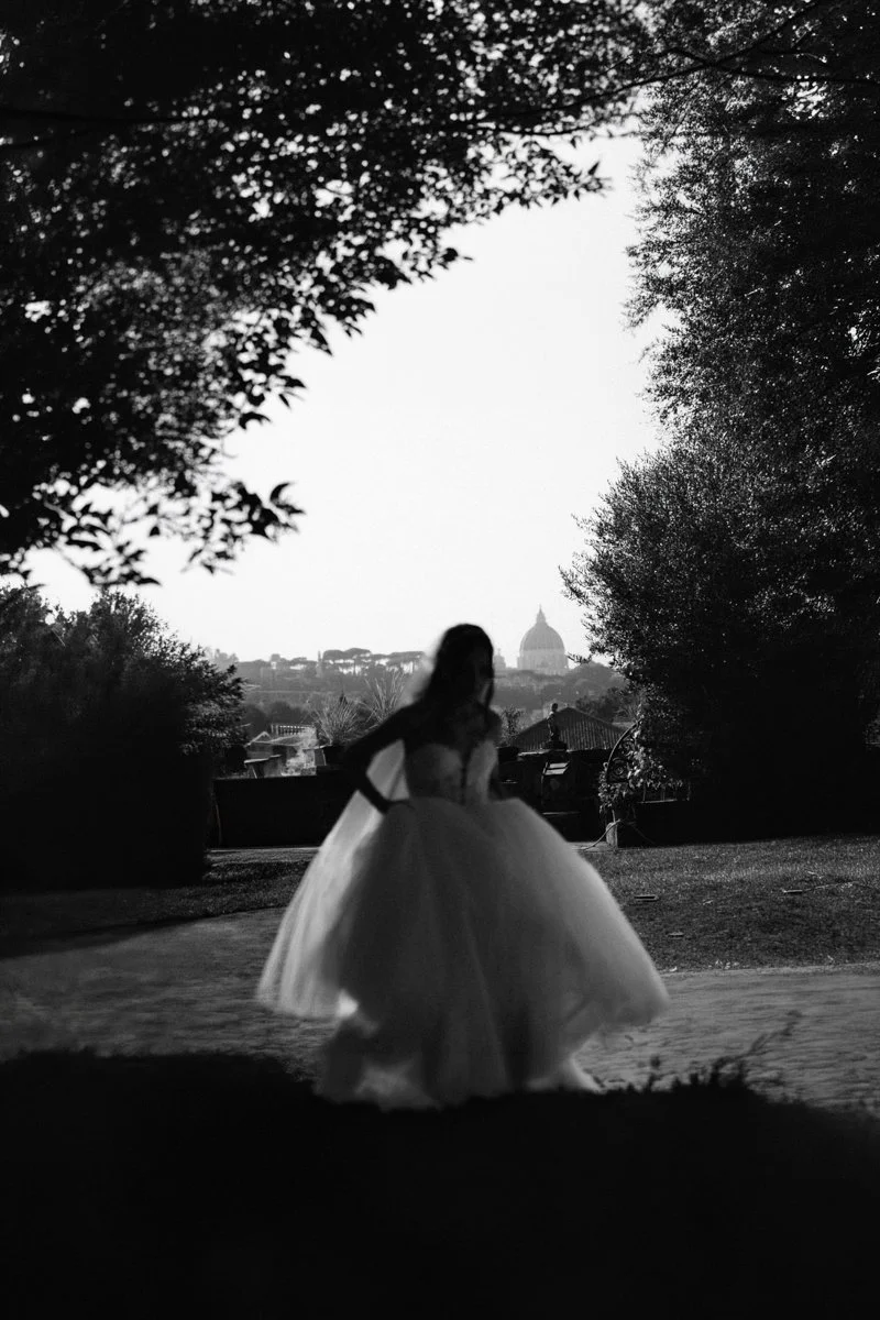 A woman in a wedding dress standing outdoors with the silhouette of a cityscape, including the dome of a prominent building, in the background during sunset or dusk.