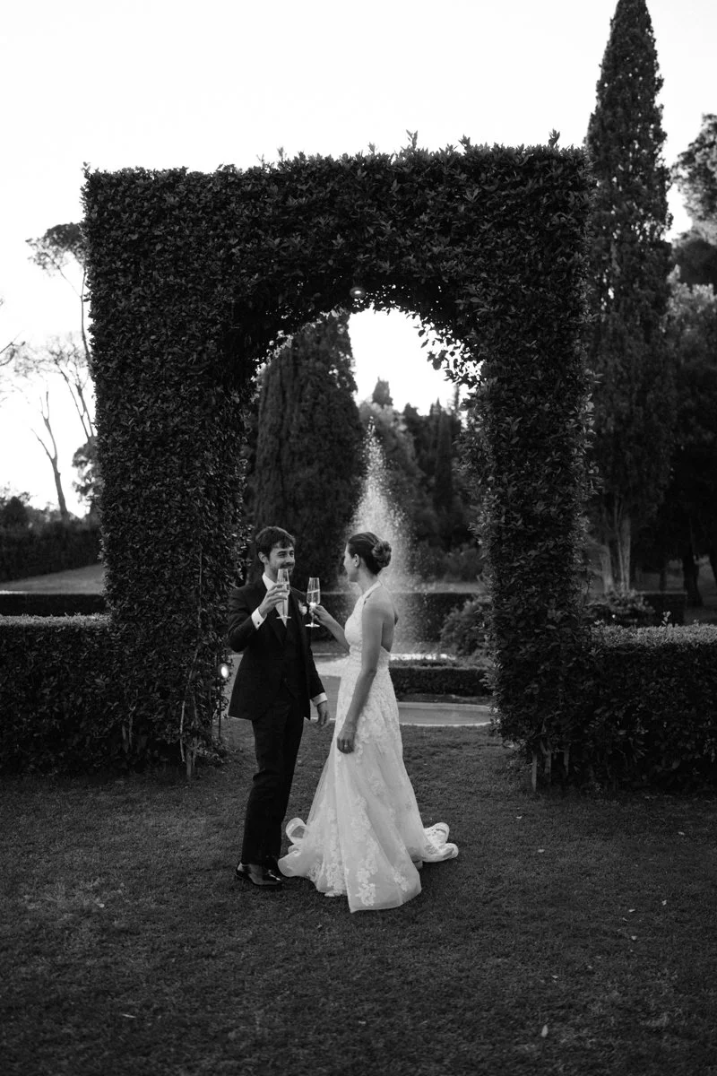 Black and white photo of a bride and groom clinking champagne glasses under an archway of dense foliage, with a fountain and trees in the background.