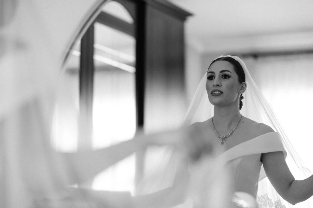 A bride in a wedding dress and veil looking at herself in a mirror.