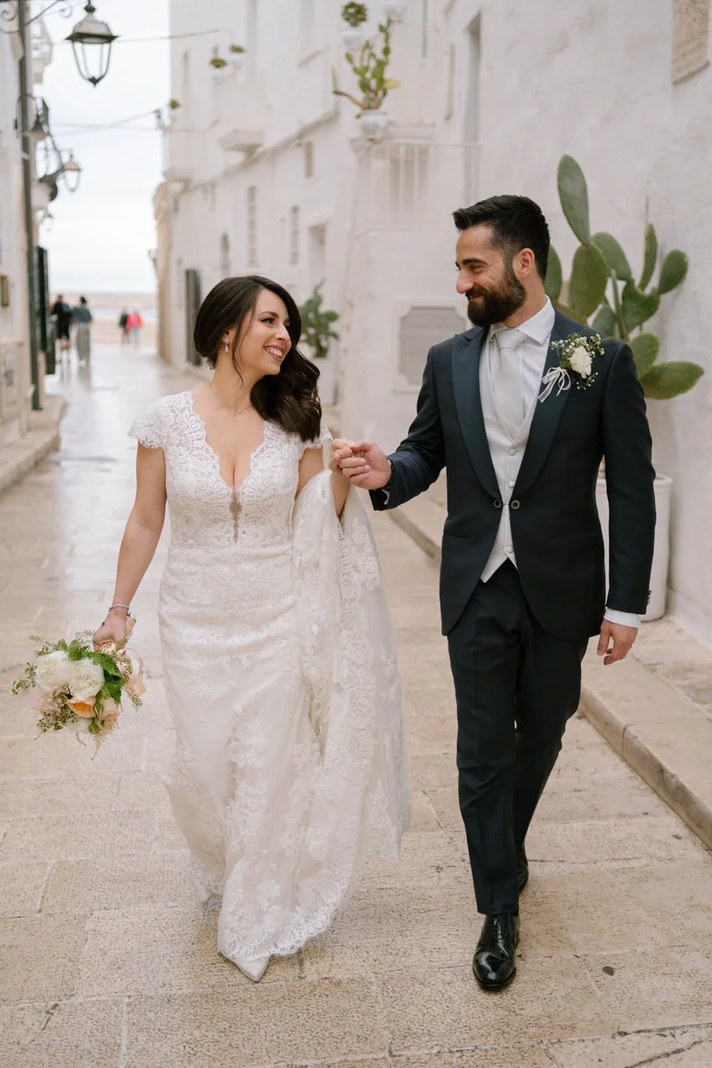 Bride and groom walking together on a street, smiling and holding hands. The bride is in a lace wedding dress holding a bouquet, and the groom is in a dark suit with a boutonnière, both looking at each other.