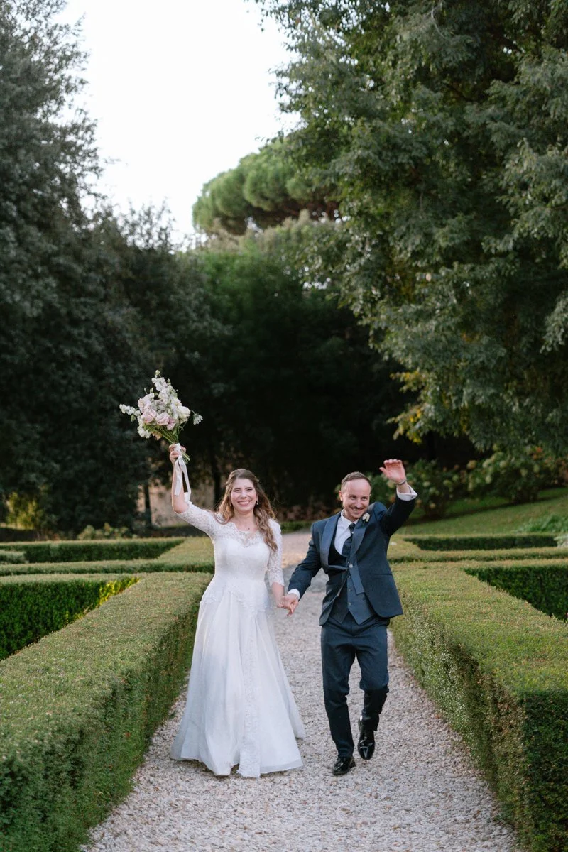 A happy couple in wedding attire walking on a garden path, smiling and waving, with the bride holding a bouquet of flowers.