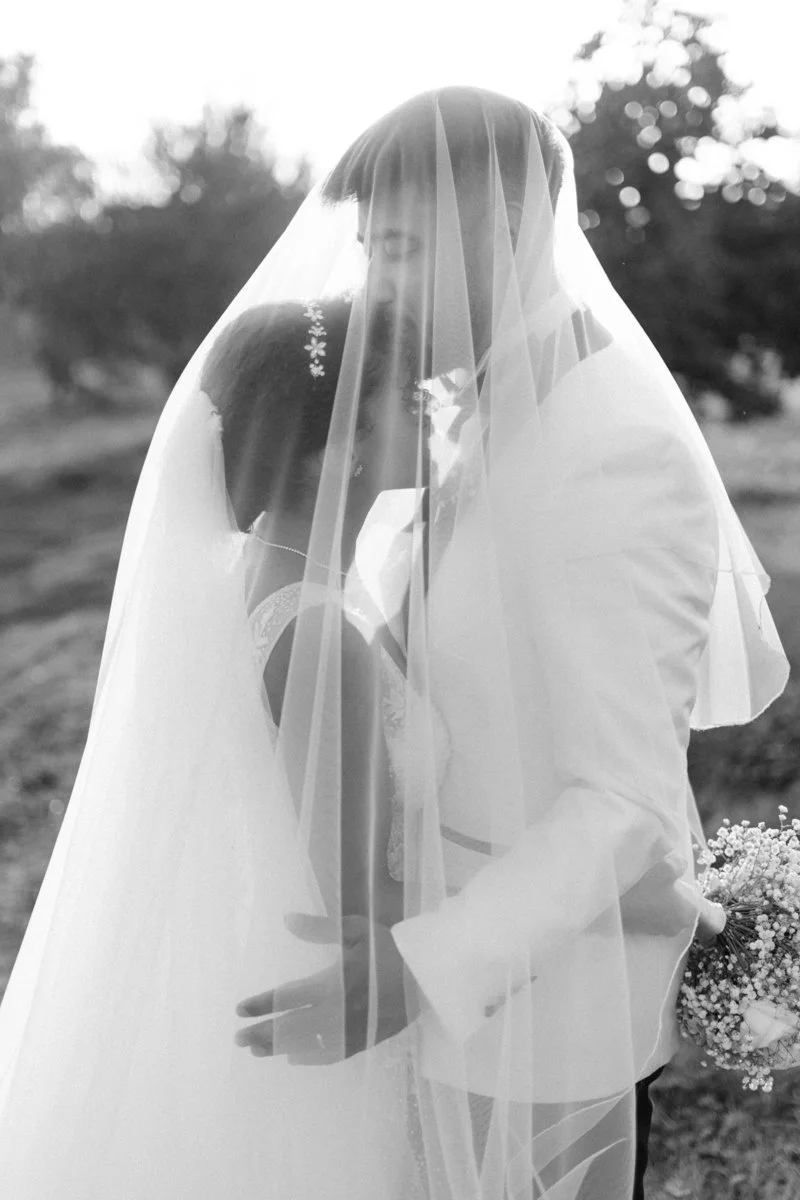 A bride and groom sharing a kiss beneath a veil on their wedding day outside with trees and sunlight in the background.