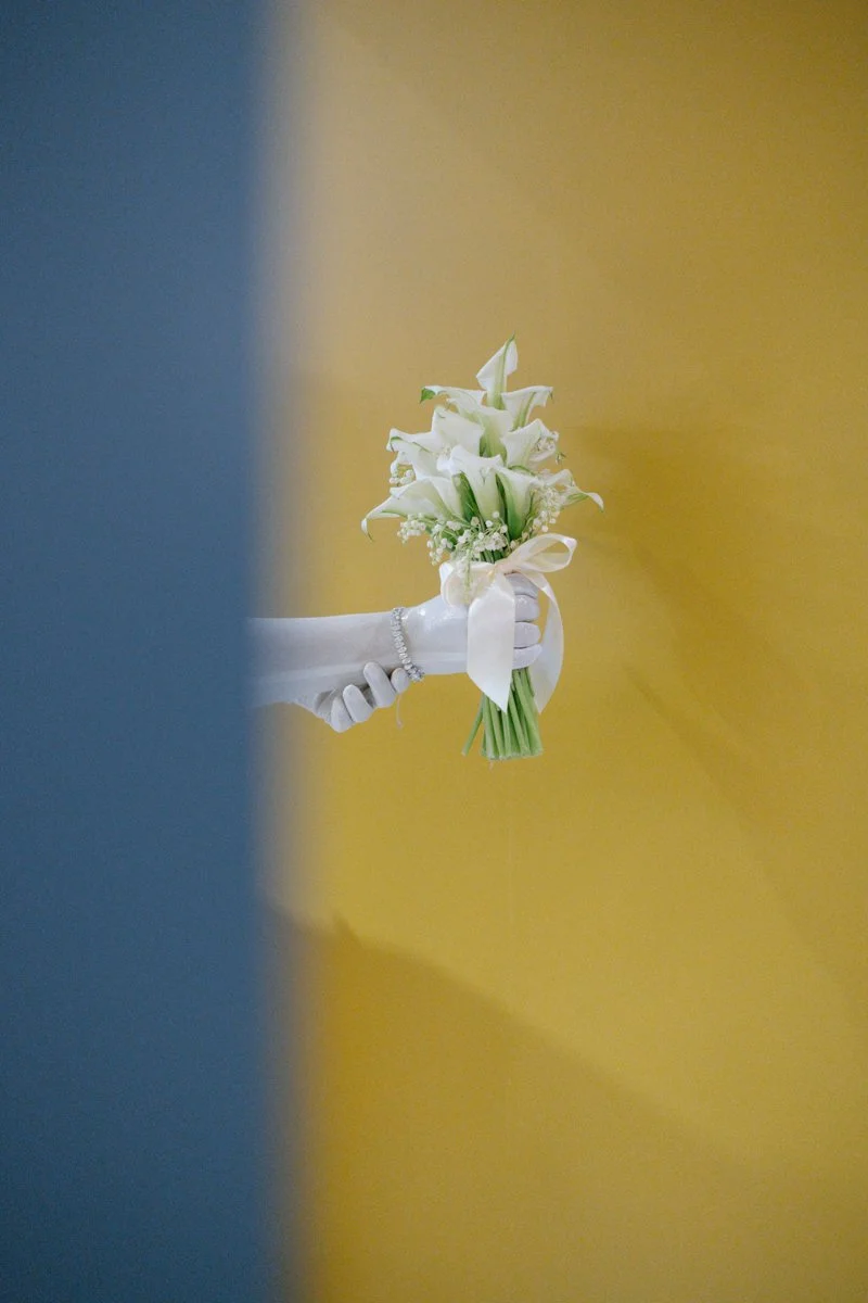 A person holding a bouquet of white calla lilies and baby's breath against a yellow background, partially obscured by a blue object in the foreground.