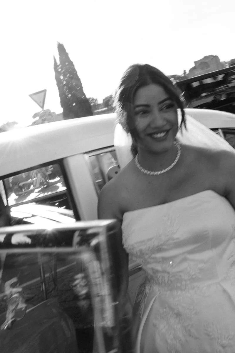 A woman in a white strapless dress and pearl necklace smiling at a celebration or event, with a car and a tall, pointed structure in the background.