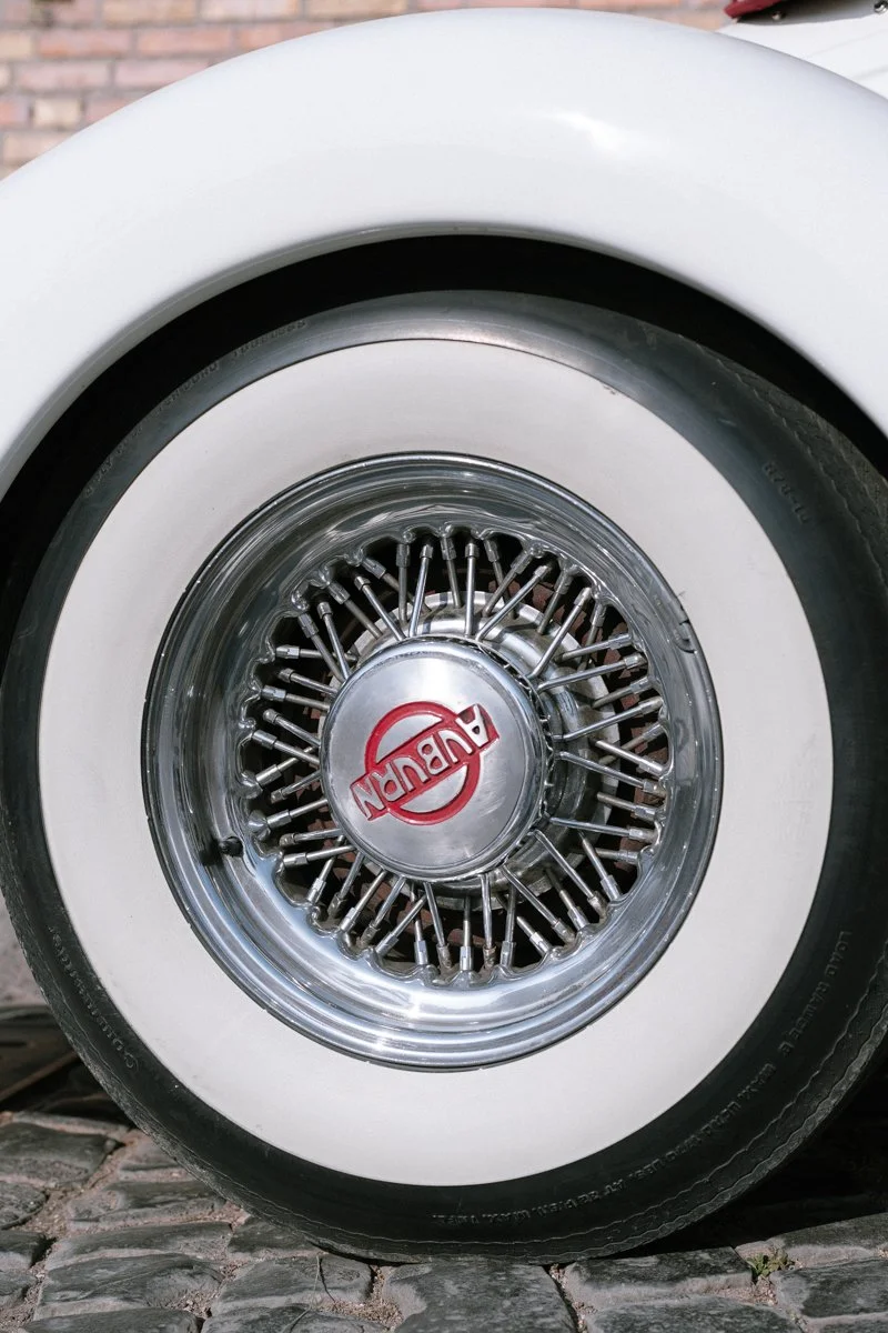 Close-up of a vintage car wheel with wire spokes, whitewall tire, and hubcap featuring the AUBURN logo, parked on cobblestone pavement.