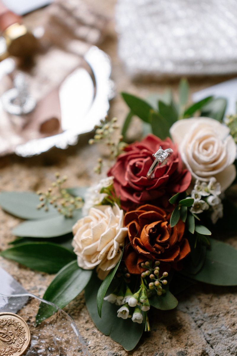 A close-up of a wedding bouquet with white, red, and brown roses, green leaves, and small white flowers, featuring a diamond engagement ring placed on a red rose.