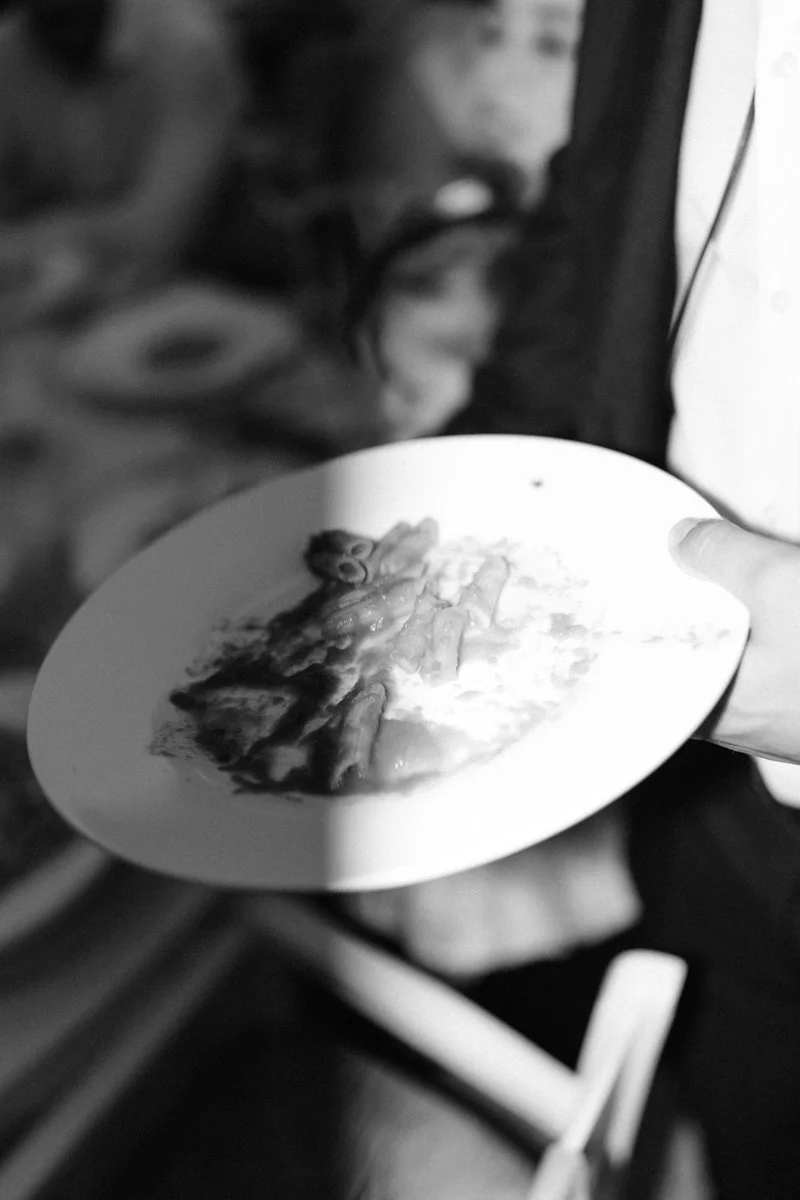 A person holding a plate with mashed potatoes and gravy, with a blurred background of elderly woman and kitchen items in black and white.