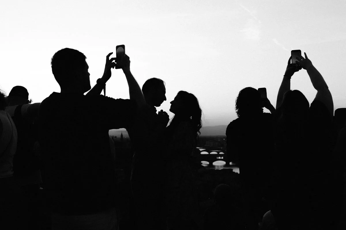 Silhouettes of people taking photos and looking at each other during sunset with a bridge and river in the background.