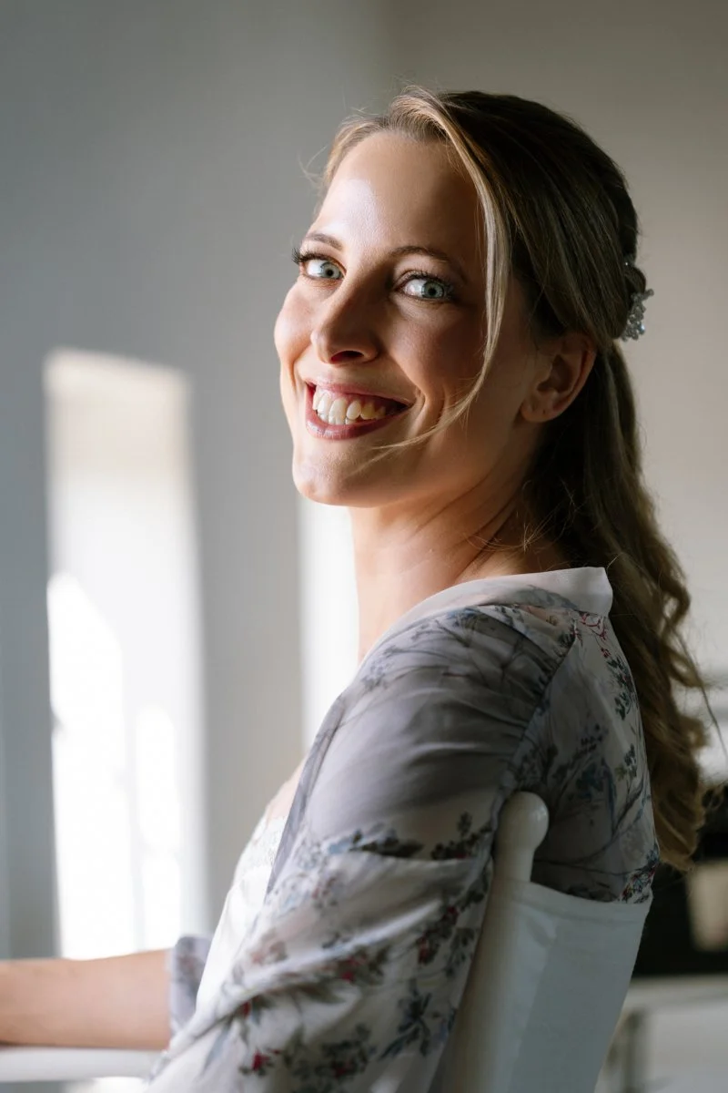 Smiling woman with blonde hair and blue eyes looking at the camera, sitting indoors near a window.