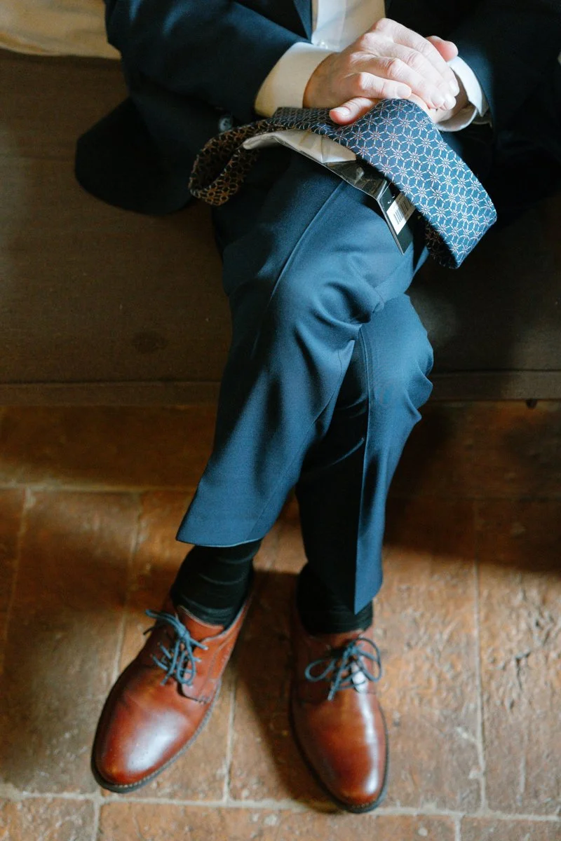 A person dressed in business attire sitting on a wooden bench, with crossed legs, holding a patterned tie in their hands, wearing brown dress shoes, dark trousers, and a dark blazer.