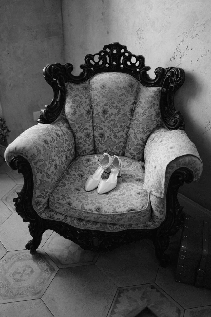 An ornate vintage armchair with patterned upholstery and dark carved wood frame, placed on a tiled floor against a plain wall. A pair of white high-heeled shoes rest on the seat.