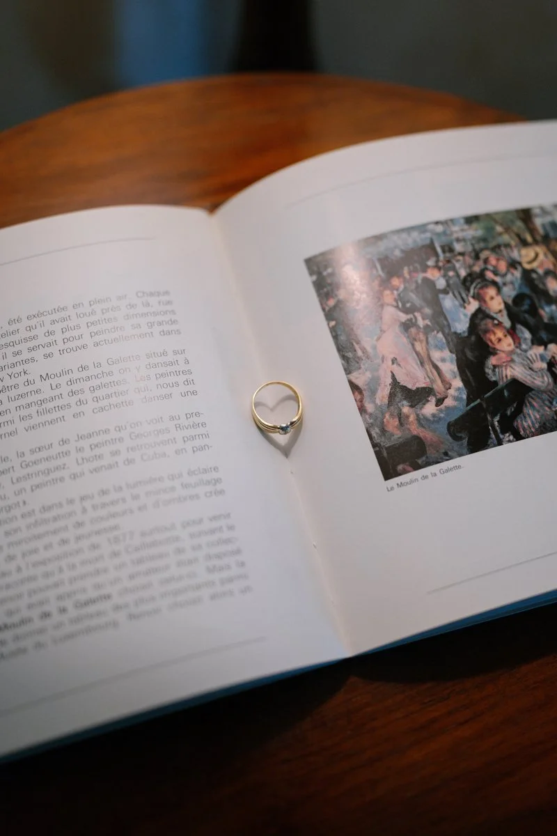 An open book with French text and a painting of a crowded scene with dancing people on the right page. A silver ring with a diamond is placed on the left page.