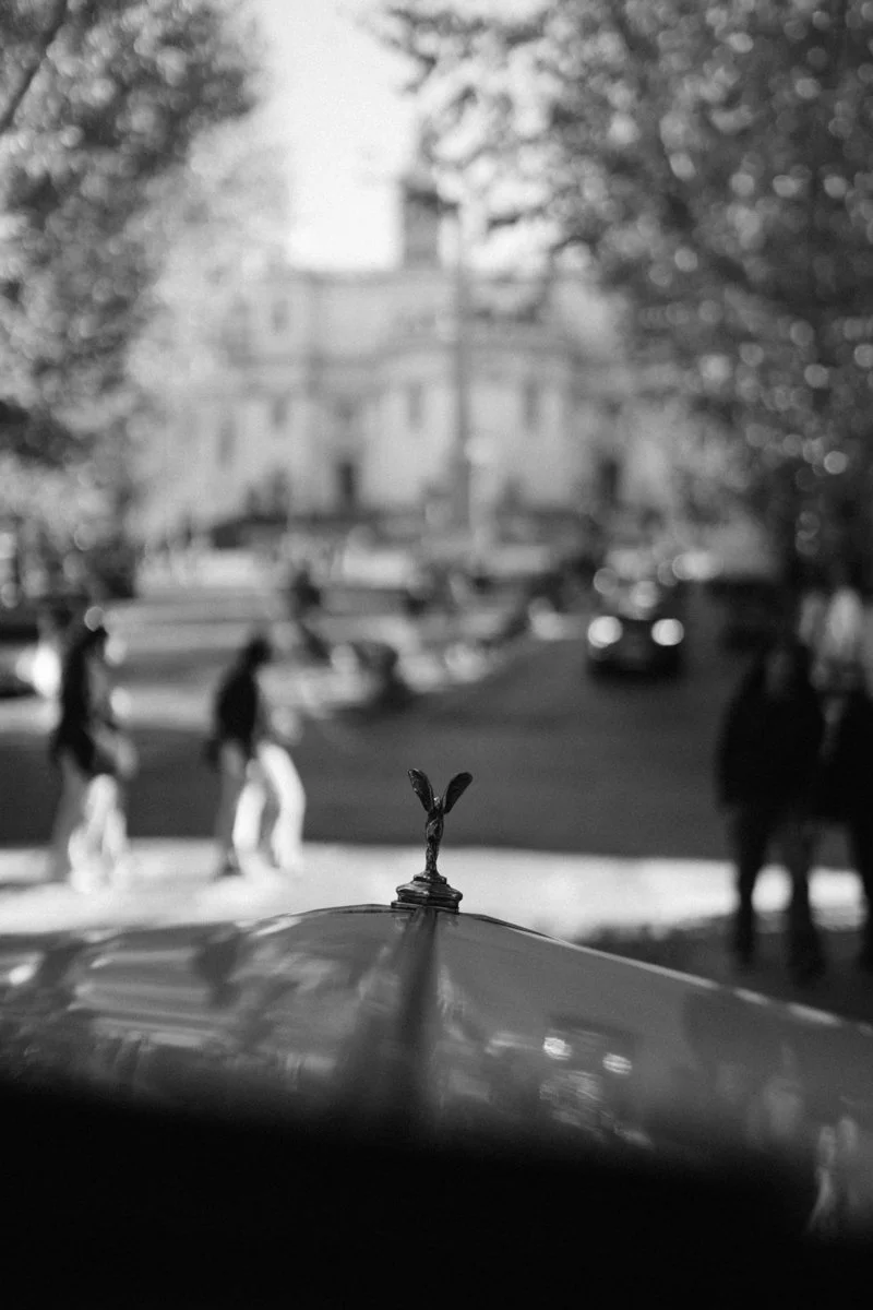 Close-up of the hood ornament on a car, with a city street scene in the background featuring pedestrians and trees.