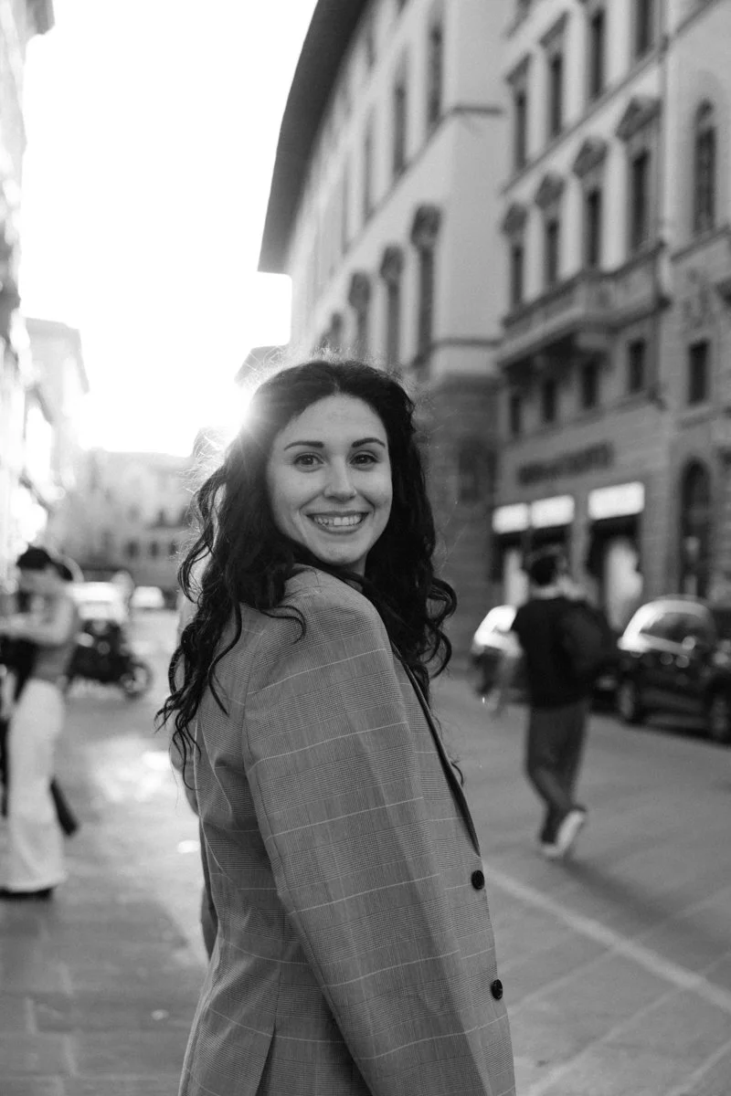 A smiling woman with dark, curly hair walking on a city street at sunset in black and white.