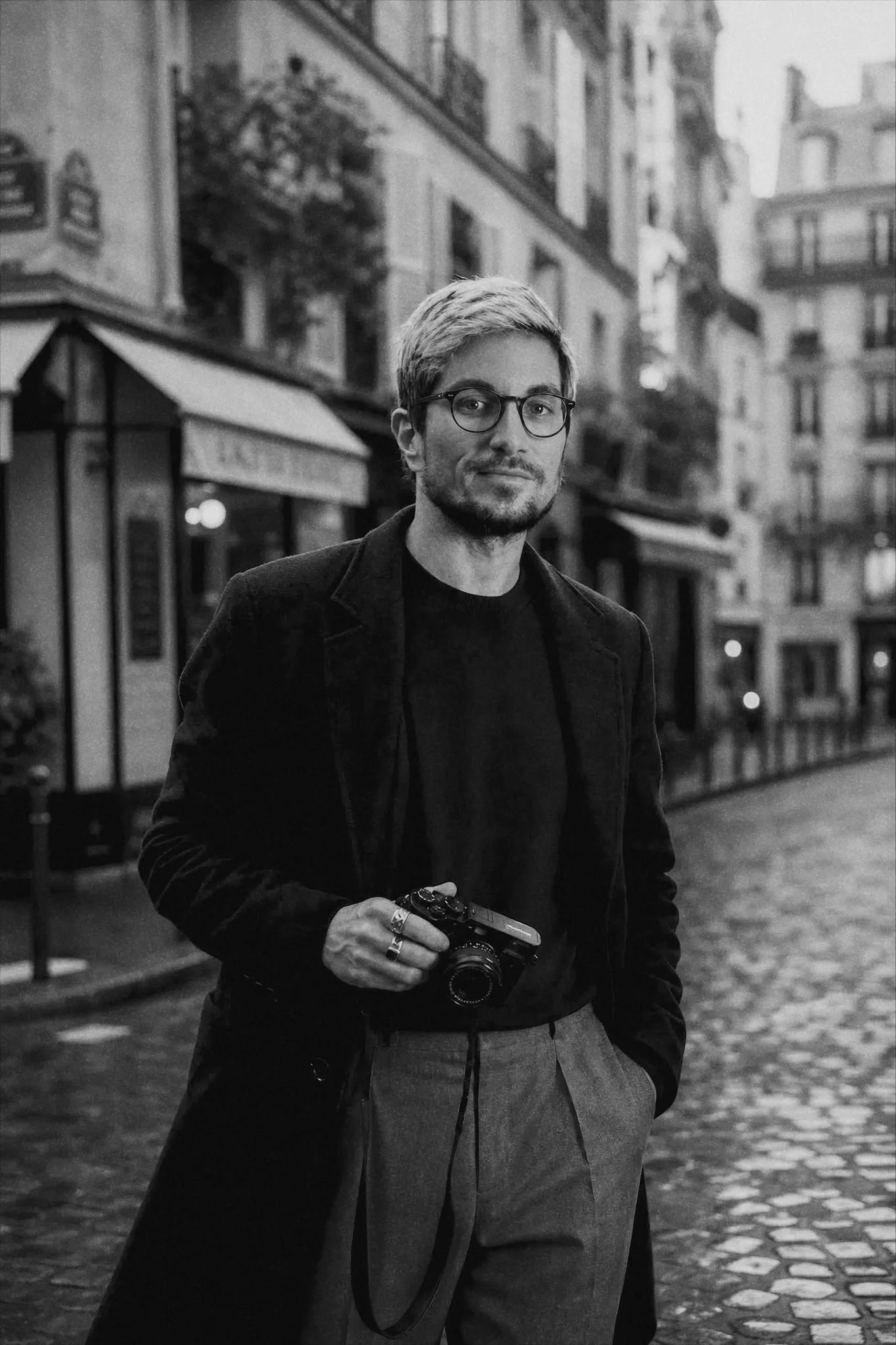 A man with glasses holding a camera, standing on a wet cobblestone street in an urban area with tall buildings and shops in the background, in black and white.