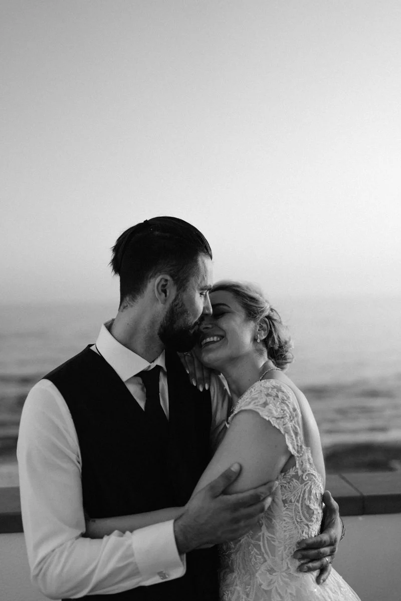 A black-and-white photo of a couple in wedding attire embracing and smiling on a balcony by the ocean, with the man gently touching the woman's arm and their foreheads touching.