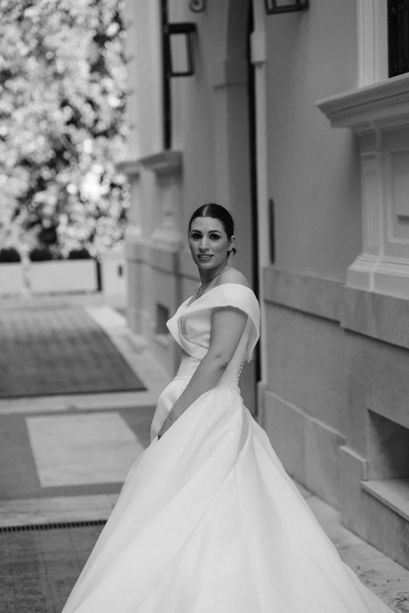 A woman in a white wedding dress standing outside a building, smiling at the camera.