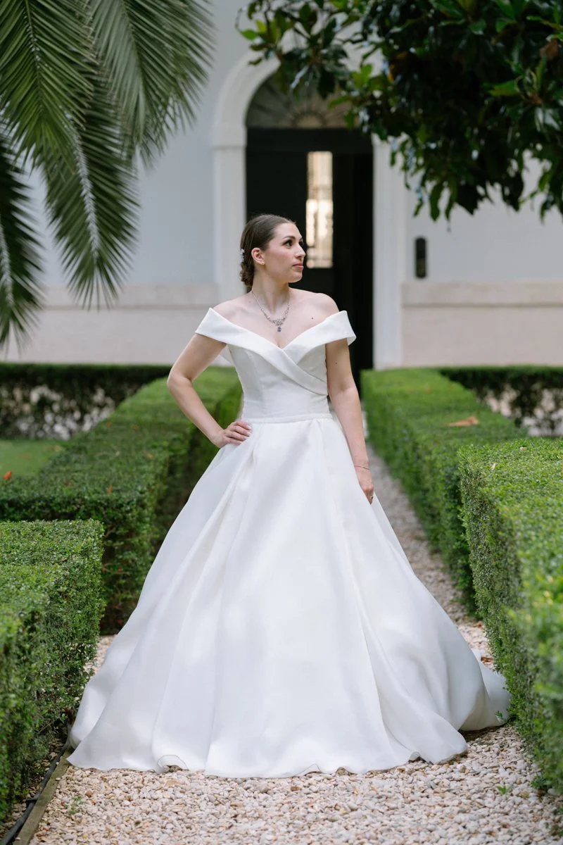A woman in a white wedding dress standing on a gravel path surrounded by neatly trimmed bushes and greenery, with a building entrance in the background.