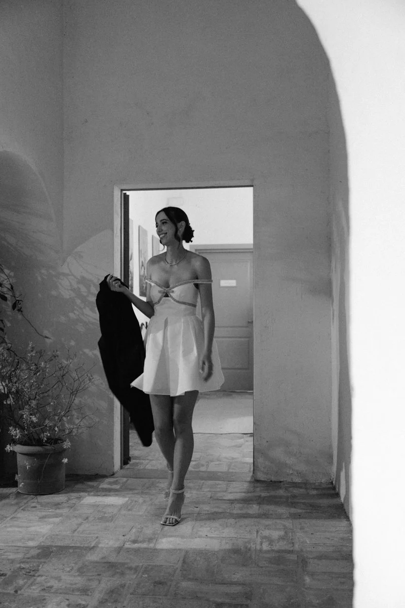 A smiling woman in a light-colored dress and high heels walking through an open doorway, holding a dark jacket in her right hand.