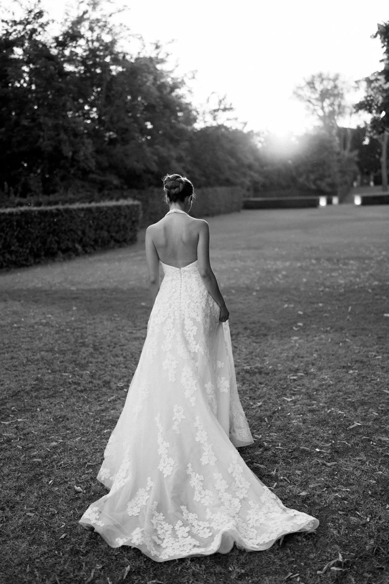 A woman in a strapless, backless wedding gown walking on a grassy area outdoors in black and white.